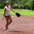 A woman in a white sleeveless top and dark shorts throws a ball while holding a black glove, standing on a dirt field with green grass and trees in the background.