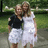 Two people stand close together outdoors; one wears a black top and white shorts, the other a white dress, with green foliage and a blurred background.