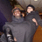 Two figures in gray hooded garments embrace against a blurred background with dark and brown tones.
