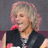 Pixelated image of a person with light-colored hair wearing dark clothing and red accents, positioned against a muted background.