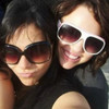 Two people wearing sunglasses pose closely together in a selfie-style image with dark hair and dark clothing.