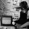 Black-and-white image of a person sitting on a newspaper-covered floor, holding a television set with a static screen.