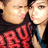 Two people in close proximity with one person's face near the other's cheek, wearing dark and red clothing against a light background.