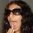 Pixelated static image of a person with long dark hair wearing black sunglasses, light skin tone, and pink lips in profile view against gray background.