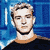 Pixelated static image of a person with light brown hair and facial stubble against a blue background, wearing a dark shirt with an orange collar.