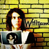 A person with shoulder-length dark hair stands against a yellow brick wall holding a small framed photo of themselves, with partial text reading 'William' on the wall.