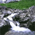 Static pixelated image showing a stream flowing over rocks with greenery in the background.