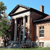 Pixelated image of a two-story house with columns, a chimney, and a tree on the left under a blue sky.