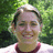 Pixelated static image of a person with light skin and dark hair smiling, wearing a maroon and white shirt against a green foliage background.