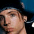 Pixelated close-up of a person's face with light skin tone, visible ear, and dark hair against a dark blue background.