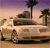 A light-colored sedan parked on a sandy surface under a gradient sky with palm trees and rocky formations in the background.