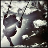 A grayscale image of a dark-colored swing suspended by two ropes against a cloudy sky background with scattered clouds.