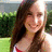 A pixelated static image shows a person with long brown hair wearing a red and white top, standing outdoors with green foliage and a light-colored structure in the background.