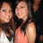 Two people with long dark hair pose closely together against a black background, one wearing a white top and the other in a sleeveless coral-colored garment.