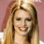 A pixelated close-up of a person with long light brown hair, smiling with visible teeth against a pinkish background.
