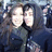 Two people in dark hooded garments pose closely together, smiling, with a blurred background of indistinct figures.