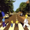 Four yellow-haired cartoon characters stand on a crosswalk in a suburban street scene with trees and cars.