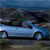 A light blue sedan with dark windows and silver wheels is positioned on a grassy area under a blue sky, with blurred trees in the background.