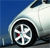 Pixelated close-up of a white car wheel with a silver multi-spoke rim, black tire, and small red detail on the rim, set against a solid blue background.