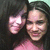 Two people in a close-up pose with one person's head tilted toward the other, wearing pink tops against a light green background.
