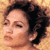 Static pixelated image of a person's face with curly dark hair against a light brown background, rendered in low resolution with visible dithering and color blending.