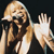 Pixelated image of a person with light brown hair holding a microphone, raising a hand in a waving gesture against a dark background.