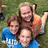 Three people pose together outdoors; two wear blue shirts with white text, one wears an orange shirt, all have brown hair and beards, background is green grass.