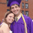 Two people in purple graduation gowns with yellow stoles stand close together against a brick building background.