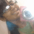 A person with dark hair and glasses holds a pink and silver camera, wearing a blue and green sleeveless top, with a bright background.
