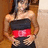 Pixelated image of a person with long dark hair wearing a black top with red horizontal stripe and black bottoms, standing against a brown and beige background.