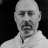 Pixelated grayscale portrait of a bald man with short facial hair, wearing a light-colored collared shirt against a solid black background.