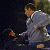 A pixelated figure in a light gray jacket and dark pants stands against a dark background with blurred green and brown elements, holding a small orange object.