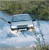 A pixelated static image shows a light blue vehicle on a reflective surface with green foliage in the background.