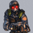 Pixelated figure in dark tactical gear with red glowing eyes, green shoulder accents, and an orange circular mark on the right arm, set against a light gray background.