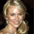Static pixelated image of a person with light brown hair styled in an updo, wearing a light-colored sleeveless top and a small earring, against a dark background.