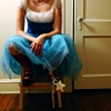 A person in a blue and white dress sits on a wooden chair with hands clasped, wearing striped socks and a star-shaped object attached to the chair.
