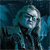 Pixelated image of a person with light hair and dark clothing against a blue-toned background, showing partial facial features and a dark collar.