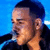 Pixelated close-up of a person with short dark hair against a blue background, wearing dark clothing and holding a microphone near the mouth.