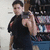 A person in a black sleeveless shirt holds a camera, positioned in front of a bookshelf with colorful spines.