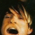 Pixelated close-up of a person's face with light brown hair, mouth open in an expressive shout against dark background.