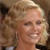 A pixelated close-up of a smiling person with light-colored hair, wearing a dark top against a blurred background.