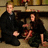 Two figures in dark formal attire sit side-by-side on a bench with a blurred background featuring warm tones and indistinct shapes.