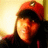 Pixelated close-up of a person wearing a red cap with a small circular emblem, dark hair, and dark clothing against a bright yellow background.