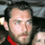 Pixelated close-up of a person with short dark hair, light skin, and blue eyes, wearing a red garment against a dark background with light patches.