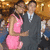 Two people in formal attire pose together against a blurred background with warm lighting.
