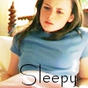 A person with long brown hair wearing a light blue t-shirt sits on a light-colored couch with a wooden headboard visible in the background.