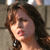 Pixelated close-up of a person with brown hair and dark eyes, mouth open in a speaking expression, wearing a dark necklace with a light pendant.
