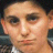 Pixelated close-up of a person's face with light skin tone, dark eyes, and visible ear, wearing a dark jacket over a light shirt.