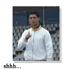A person in a white zip-up jacket with yellow trim stands against a blurred stadium background, making a shushing gesture with one hand near their mouth.