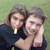 Two people in dark clothing embrace on a grassy background, with one resting their head on the other's shoulder.